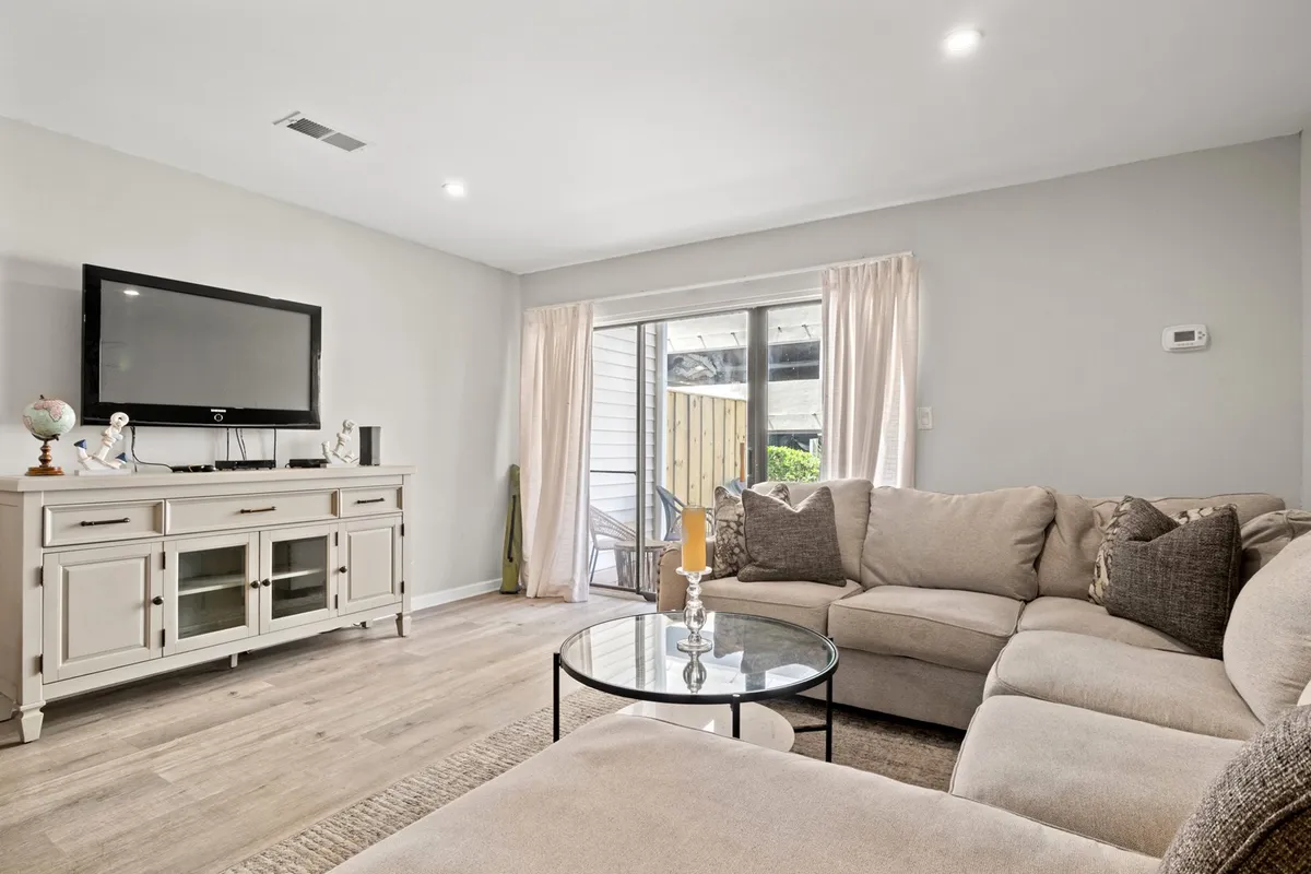 A cozy living area anchored by a large sectional sofa, a coffee table, a television, and a sliding door with curtains. The arrangement suggests a space ideal for movie nights, socializing, and relaxing after a day at the beach.