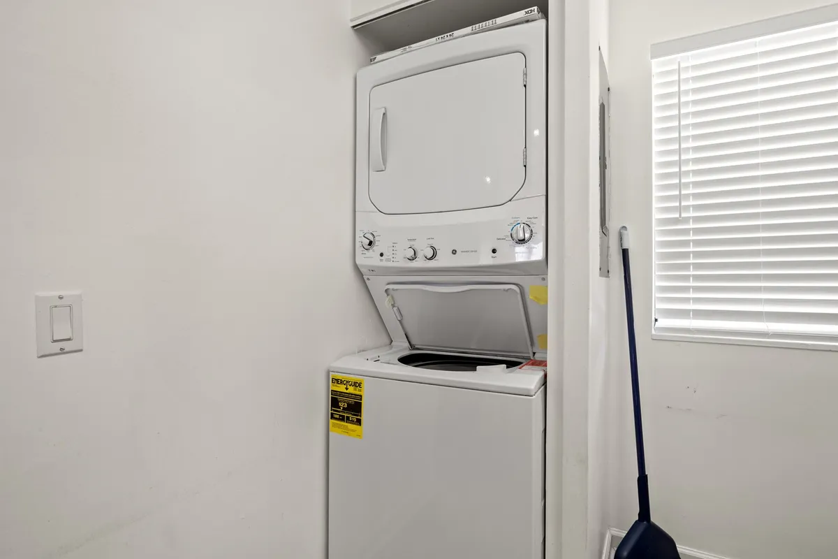 A bright and tidy laundry area with a stacked washer and dryer set beside a window with blinds, set against clean white walls. The space feels practical, organized, and easy to use.