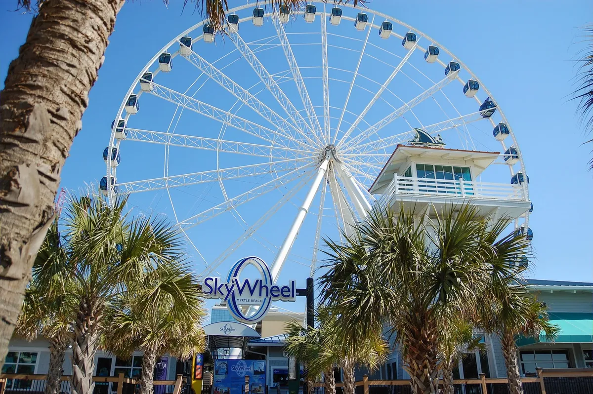 This image captures the iconic SkyWheel, framed by lush palm trees and the ticket booth below. The Ferris wheel is a vibrant focal point, and the bright, cheerful mood evokes excitement and the spirit of Myrtle Beach attractions.