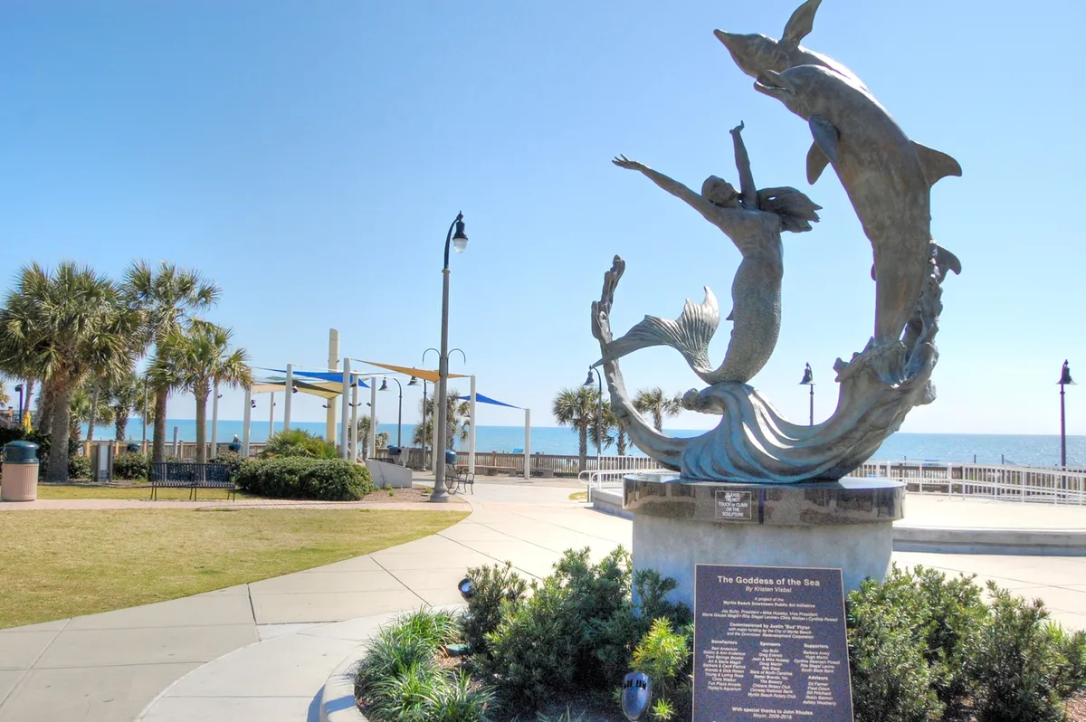 A serene beachfront scene featuring the Goddess of the Sea sculpture, palm trees, a winding walking path, and a relaxing ocean view. The setting invites visitors to explore outdoor art, take in the scenery, and create lasting memories along the Myrtle Beach coast.