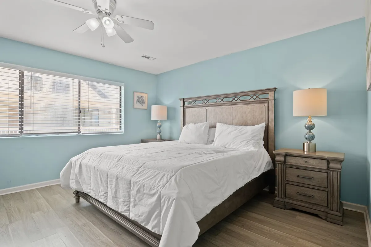 A serene bedroom scene highlights a bed with crisp white linens, flanked by wooden nightstands and decorative lamps. The large window allows natural light to flood in, enhancing the peaceful ambiance. The arrangement suggests comfort for both short and extended stays, from restful nights to sunlit mornings.