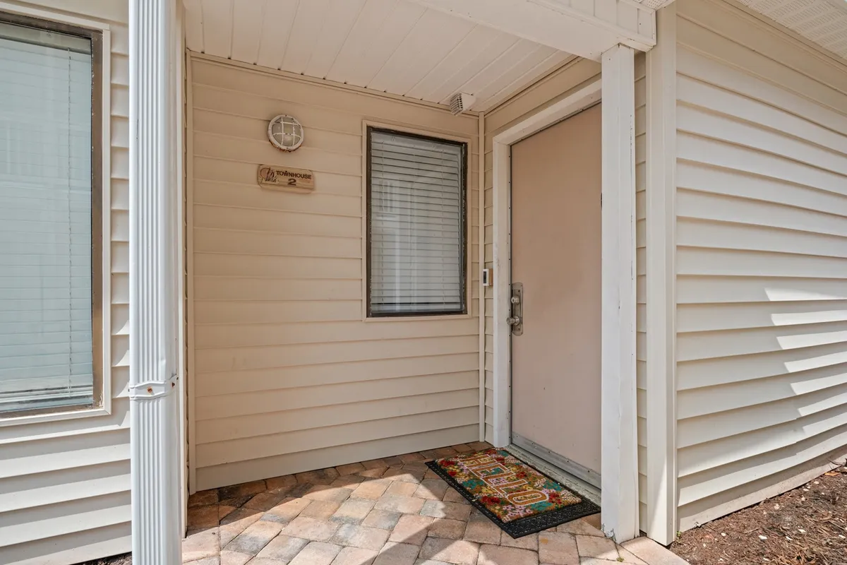 A warm and inviting interior scene focused on a decorative doormat, the front door, and a sunlit window with blinds. The space feels welcoming and homely, suggesting effortless arrivals and a comfortable stay.