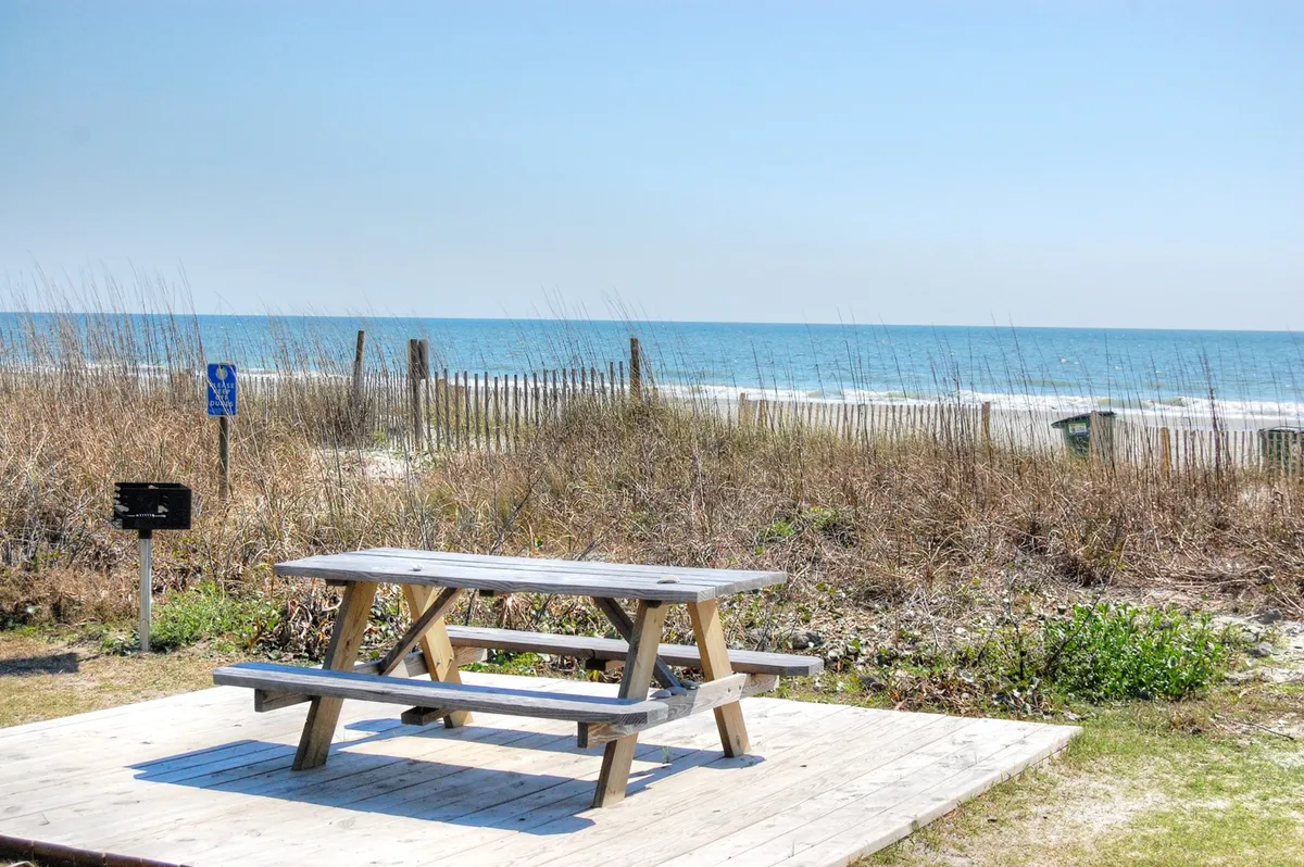A wooden picnic table is set atop dune grasses, offering an idyllic spot to savor ocean views. The expansive sea stretches into the distance, inviting guests to relax, picnic, or simply enjoy the sound of waves.