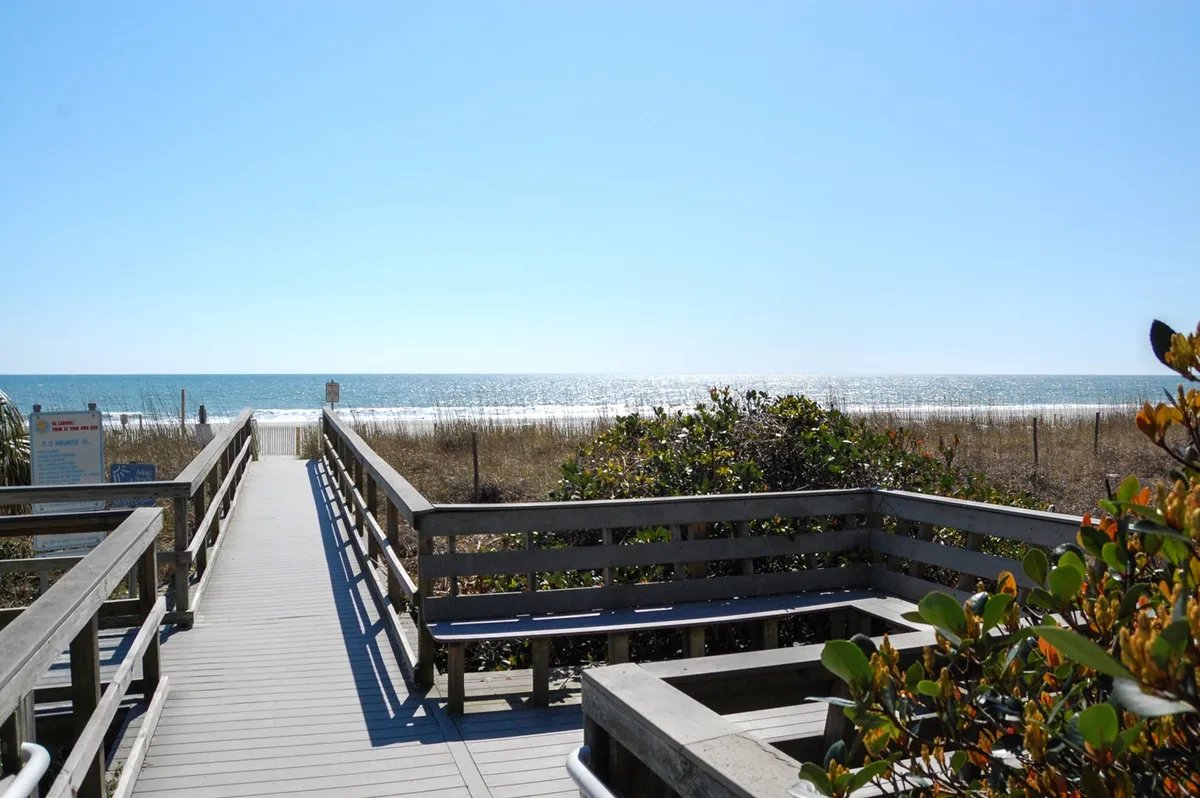 A sunlit wooden walkway stretches across dunes, guiding guests toward the sandy shoreline. The ocean glimmers in the background, with soft beach grass framing the path and distant figures enjoying the serene beach setting.