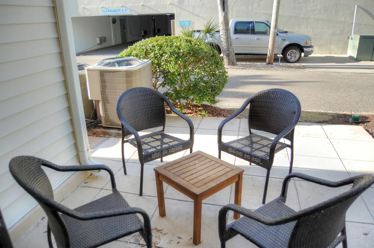 A relaxed patio setup with four outdoor chairs around a wooden table, surrounded by lush greenery. The scene suggests casual, social outdoor gatherings, such as morning coffee, sunny brunches, or evening relaxation with friends or family.