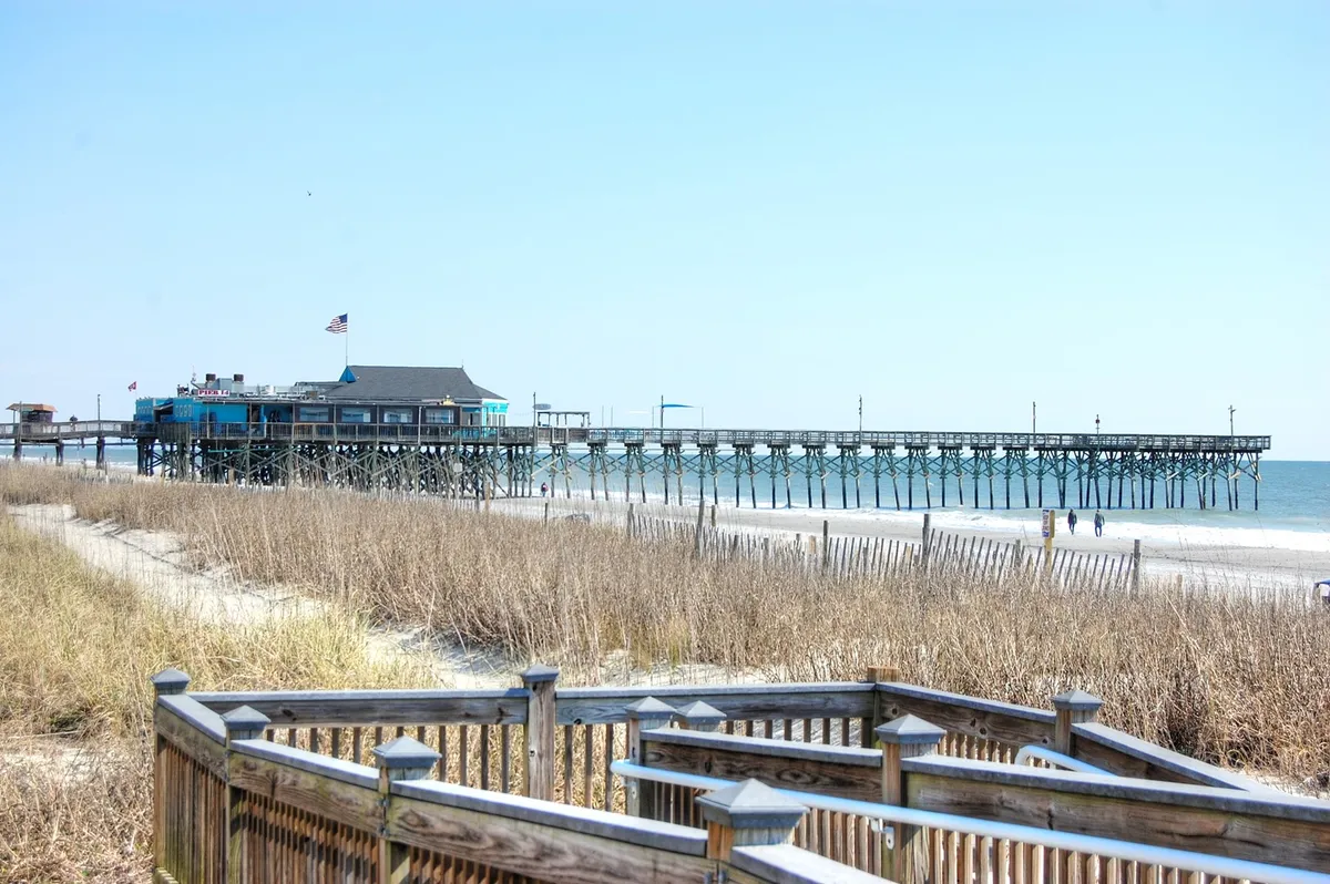 A wide view of a pier stretching out over the ocean, bordered by sandy dunes and fluttering flags. The area reflects a quintessential coastal vibe, with activities like fishing, walking, dining, or enjoying scenic sunrises and sunsets.