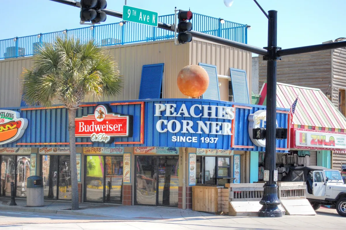 An exterior street scene featuring the iconic Peaches Corner sign alongside a Budweiser sign, colorful awnings, and hints of a lively dining atmosphere. The area appears vibrant, with people likely enjoying food and drinks outdoors and soaking up the neighborhood's energy.