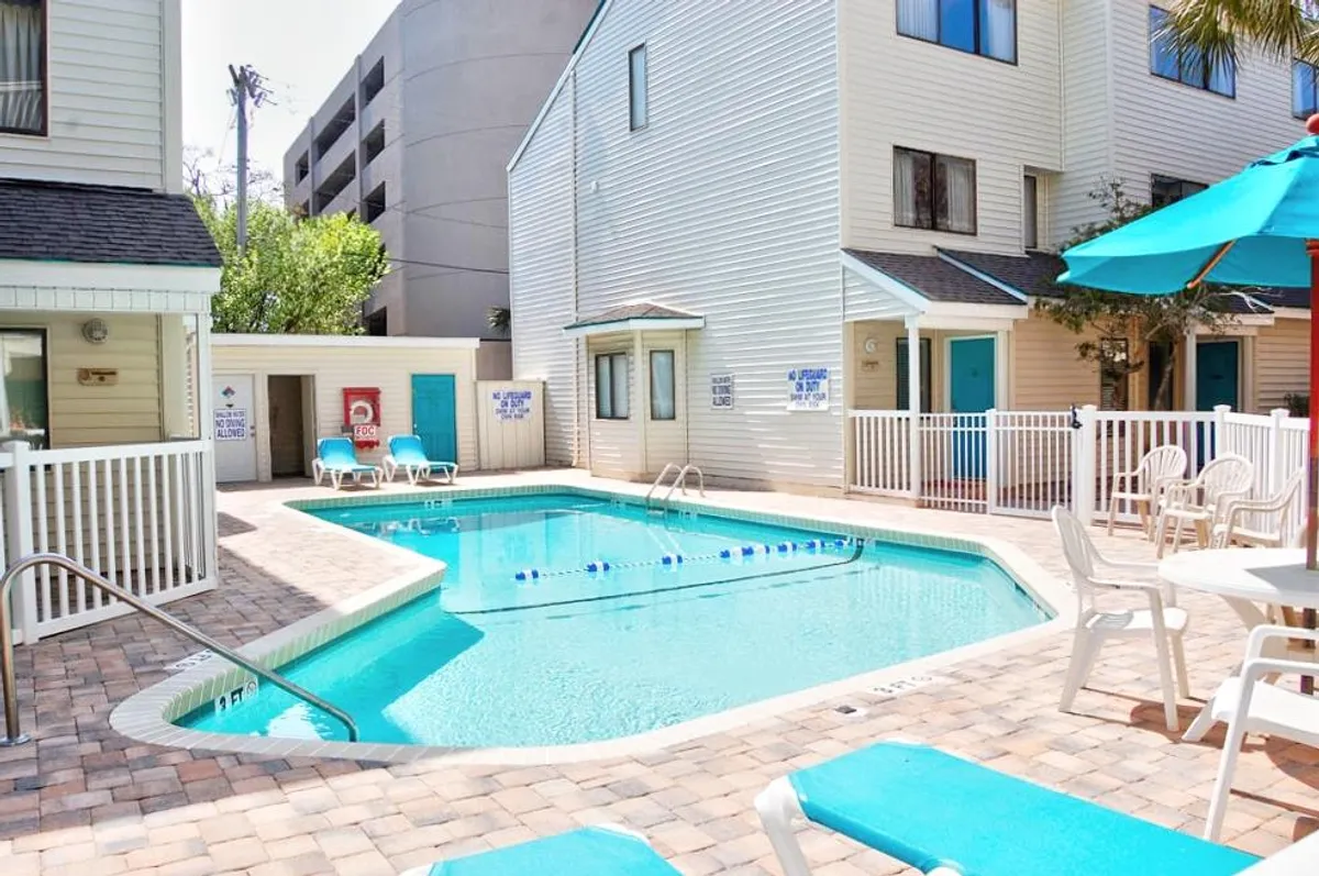The image features a sparkling outdoor swimming pool surrounded by inviting lounge chairs and colorful umbrellas, set against the backdrop of charming townhome buildings. The scene is lively and welcoming, suggesting opportunities for both relaxation and socializing in a sunny, beachside atmosphere.