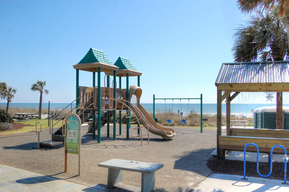 A vibrant playground with swings and slides sits in view of the sandy beach and palm trees, filled with children playing and families gathering under the sun.