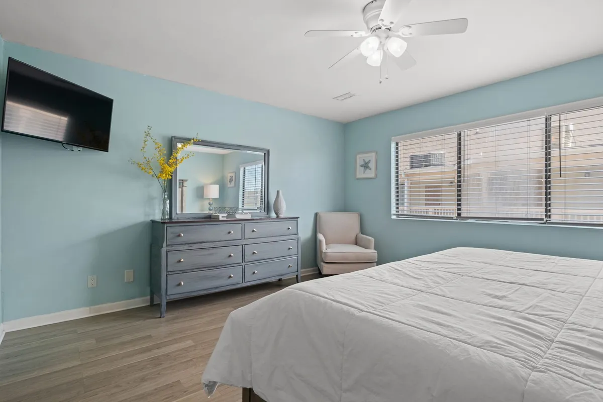 A serene bedroom setup featuring a bed, dresser, mirror, chair, and wall-mounted TV. The room is softly lit and organized for maximum comfort, offering a quiet retreat for morning coffee, afternoon reading, or relaxing movie nights.