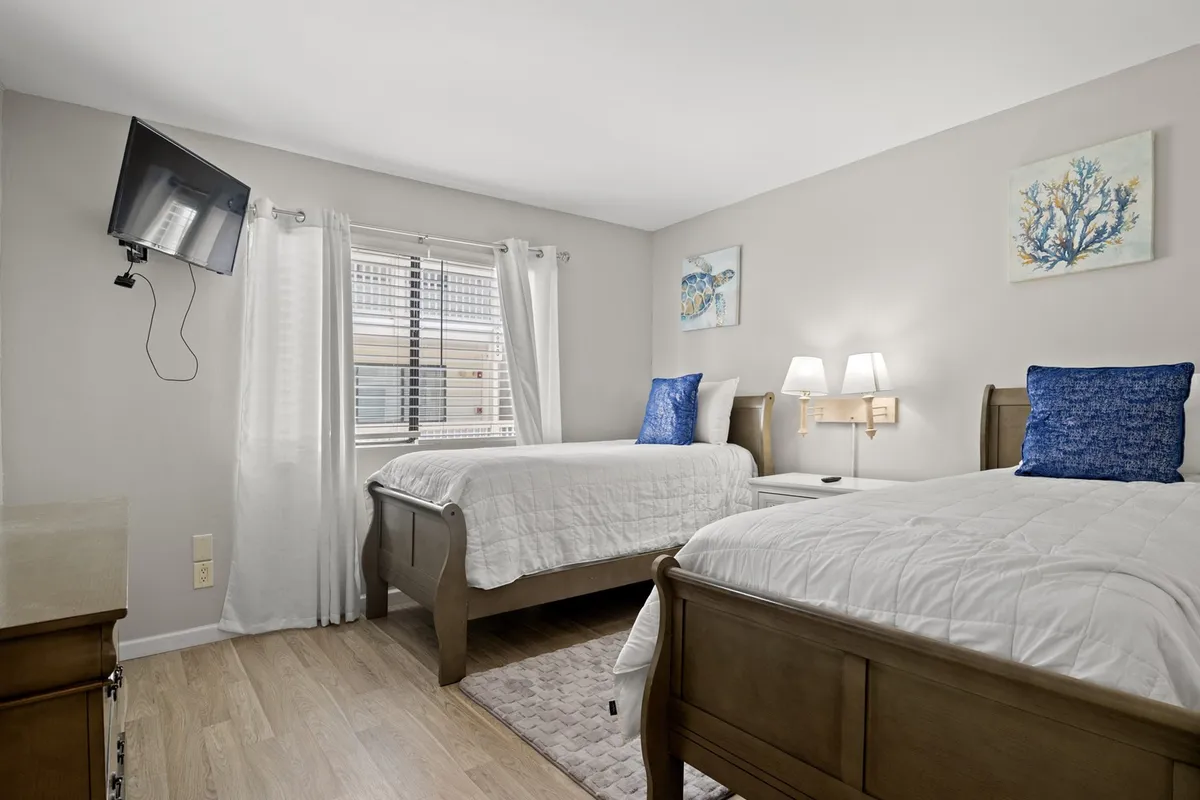 A comfortable guest room is shown with two beds, a wall-mounted TV, decorative artwork, and soft lighting from table lamps. The setup alludes to cozy movie nights, early morning coffee, and shared bedtime stories—a versatile space for families or friends traveling together.